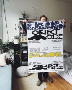 a person holding up a poster in front of a room with a couch and bookshelf