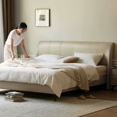 a woman in white shirt and tan pants cleaning a bed with beige linens on it