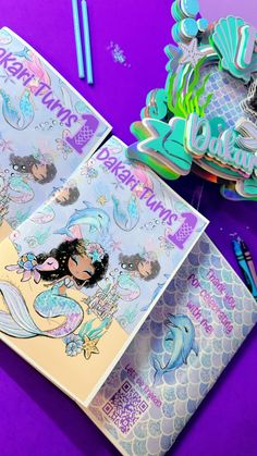 two mermaid themed birthday cards with markers and pencils on a purple surface next to them