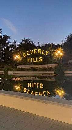 a sign that says beverly hills sits in front of a pond with lights on it