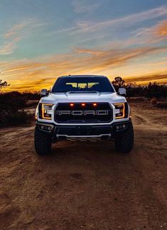 the front end of a white truck parked on top of a dirt field at sunset