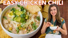 a woman holding a bowl of chicken chili next to tortilla chips and guacamole