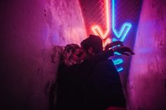 a man and woman kissing in an alleyway with neon signs on the wall behind them