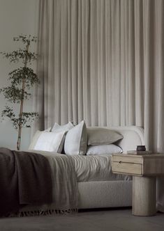 a bed with white sheets and pillows next to a small tree in front of a curtained window