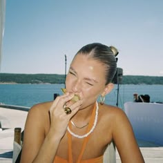 a woman sitting at a table eating food with a drink in front of her and the water behind her