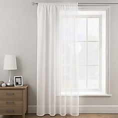 a white curtain hanging on the side of a window next to a nightstand with a lamp