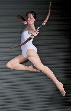 a woman in white leotard holding a bow and arrow with both hands while jumping up into the air