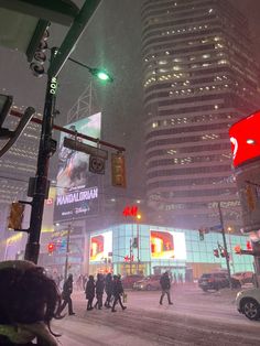 people walking in the snow at night near tall buildings and traffic lights on a city street