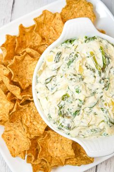 a bowl of dip surrounded by tortilla chips on a white plate with a wooden table