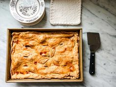 an apple pie in a baking pan next to a spatula and spoon on a marble counter top