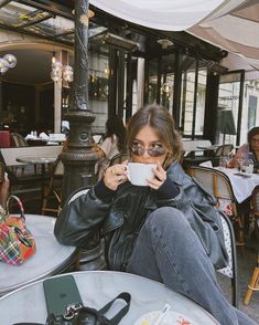 a woman sitting at an outdoor table drinking from a coffee cup and looking into the camera