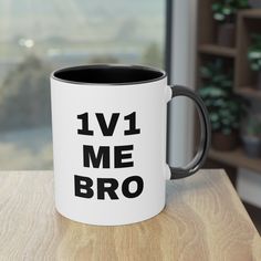 a black and white coffee mug that says 1v1 me bro sitting on a wooden table