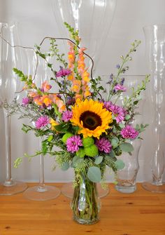 a vase filled with lots of colorful flowers on top of a wooden table next to wine glasses