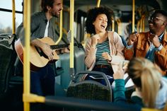 three people on a bus playing guitar and singing while another person looks at the camera