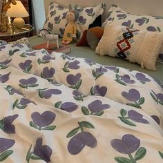 a bed with purple flowers on it next to a night stand and two stuffed animals