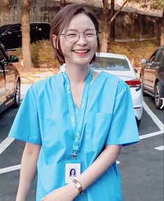 a woman in blue scrubs and glasses standing in a parking lot next to parked cars