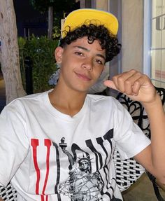 a young man with curly hair wearing a white shirt and yellow hat sitting on a bench