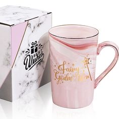 a pink coffee mug sitting next to a white and black box with gold lettering on it