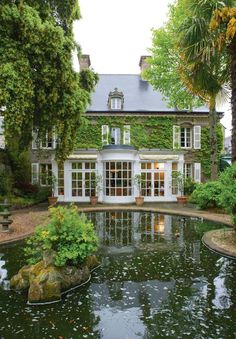 a pond in front of a large house with lots of trees and plants around it