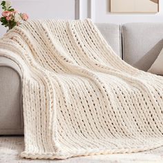 a white knitted blanket sitting on top of a couch next to a vase with flowers