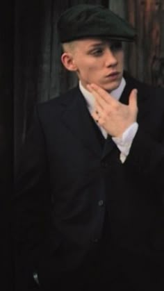 a young man in a suit and hat is holding his hand to his face while standing next to a wooden wall