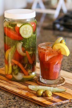 a mason jar filled with pickles, tomatoes and cucumbers next to a drink on a cutting board