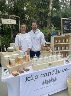 two people standing behind a table with soaps and candles on it in front of some palm trees