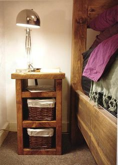 a small wooden table with baskets under it next to a bed and lamp on the wall