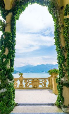 an arch covered in vines and potted plants next to the water with mountains in the background