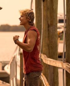 a young man standing on a dock next to the water with his hands in his pockets