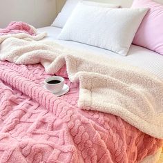 a pink blanket on top of a bed next to pillows and a cup of coffee