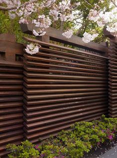 a wooden fence is lined with flowers and trees in the background, along side a sidewalk