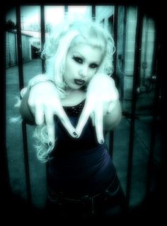 a woman with white hair and makeup holding her hands in the shape of v behind bars