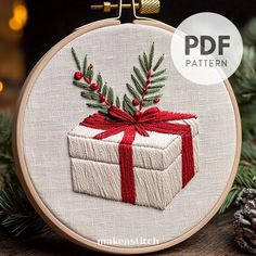 a cross stitch christmas ornament with a present on it and pine cones in the background