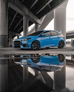 a blue car is parked in front of a bridge with its reflection on the water