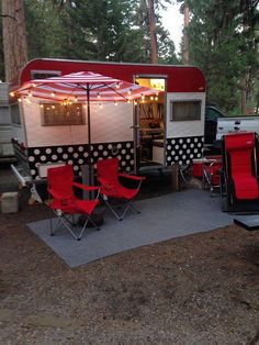 a red and white camper parked in the woods next to some chairs with umbrellas on them