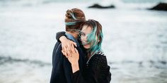 two women hugging each other on the beach by the ocean with blue hair and water in the background