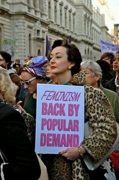 a woman holding a sign that says feminist back by popular demand on the side of her face