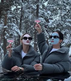 two women with face paint holding up wine glasses in the snow while sitting on top of a car