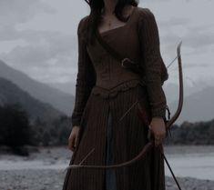 a woman holding a bow and arrow in front of a body of water with mountains in the background