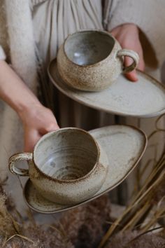 two tea cups and saucers being held by someone's hands in front of dried grass