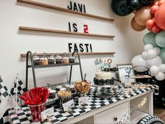 a dessert table with black and white checkered cloths on it, balloons in the background