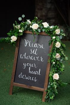 a chalkboard sign decorated with greenery and white roses is the centerpiece for an outdoor ceremony