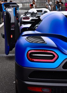 several blue sports cars are parked on the side of the road with people standing around them