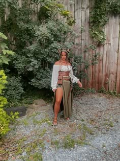 a woman wearing a skirt and top standing in front of a wooden fence with greenery