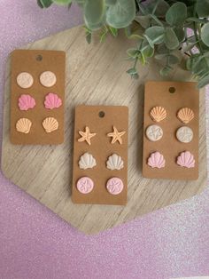 three small seashells and starfish earrings are on a wooden board next to a potted plant