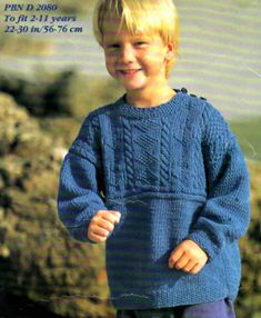 a young boy wearing a blue sweater and purple pants standing in front of some rocks