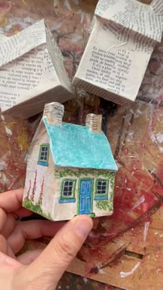 a hand holding a small house made out of paper on top of a piece of cardboard