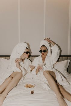 two women in white robes sitting on a bed with food and drinks, both wearing sunglasses