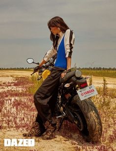 a woman standing next to a parked motorcycle in the middle of a dry grass field